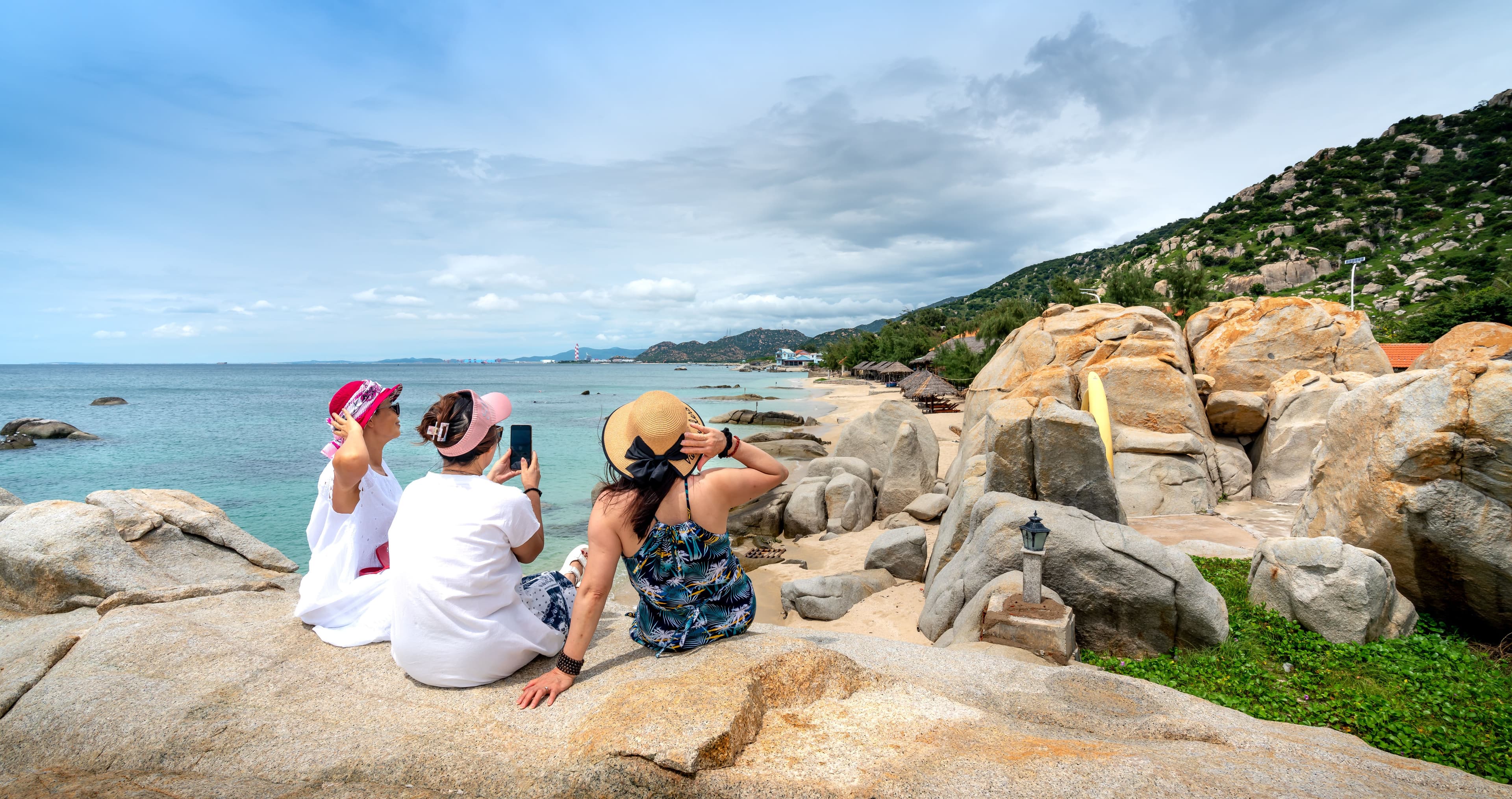 Three women sit on rocky shore capturing beach views. Scenic summer relaxation.